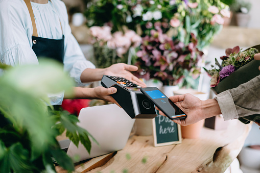 Cropped shot of young Asian man shopping at the flower shop. He is paying for a bouquet with his smartphone, scan and pay a bill on a card machine making a quick and easy contactless payment. NFC technology, tap and go concept