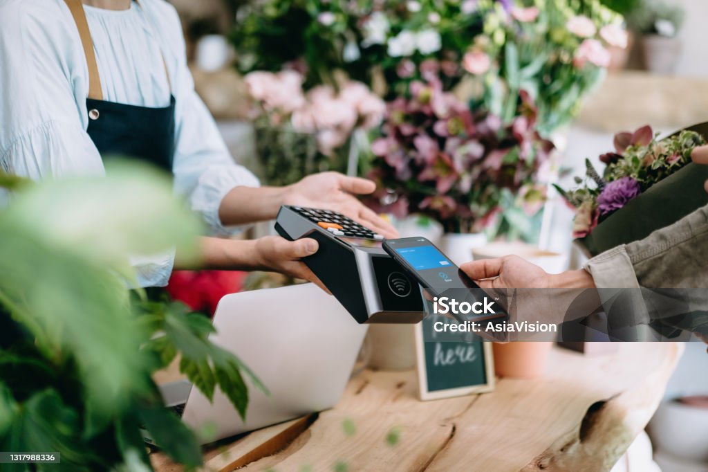 Cropped shot of young Asian man shopping at the flower shop. He is paying for a bouquet with his smartphone, scan and pay a bill on a card machine making a quick and easy contactless payment. NFC technology, tap and go concept Banking Stock Photo