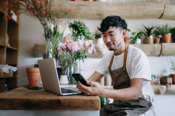 joven florista macho asiático, propietario de una floristería de pequeñas empresas, usando teléfonos inteligentes mientras trabajaba en computadoras portátiles contra flores y plantas. comprobación de stocks, toma de pedidos de clientes, venta de pro - florist small business flower shop owner fotografías e imágenes de stock