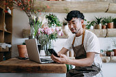 Young Asian male florist, owner of small business flower shop, using smartphone while working on laptop against flowers and plants. Checking stocks, taking customer orders, selling products online. Daily routine of running a small business with technology