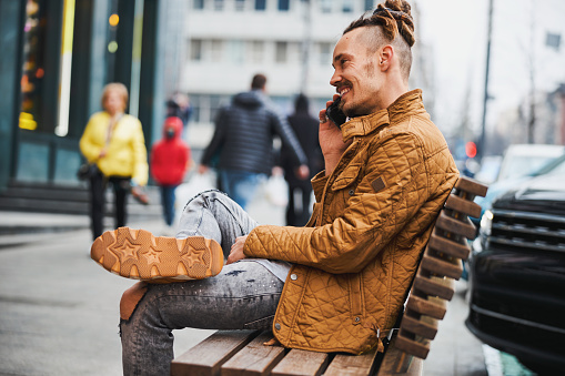 Cheerful Caucasian man sitting on a street bench with one foot on the knee and smiling during a phone talk