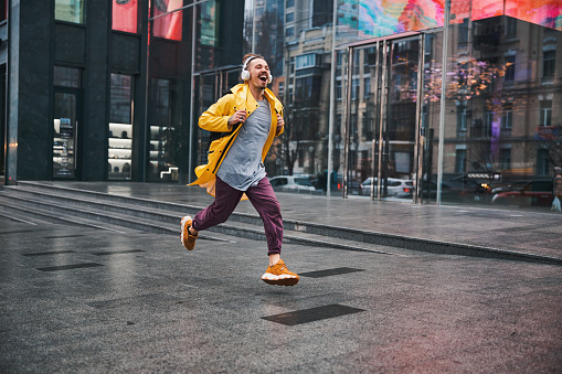 Happy trendy guy is listening to music with headphones while running along wide urban street