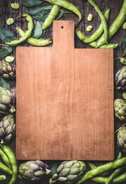Rustic wooden cooking background with vintage cutting boar, fresh  artichokes and fava beans (Fave). Springtime vegetables, ingredients of Mediterranean cuisine