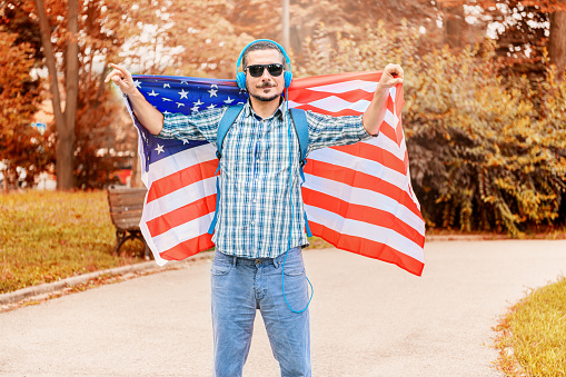 Young Tourist With Backpack is Exploring the City, Walking in the Public Park, and Eating an Ice Cream While Listening to the Music With Large Headphones and Waving With American Flag.