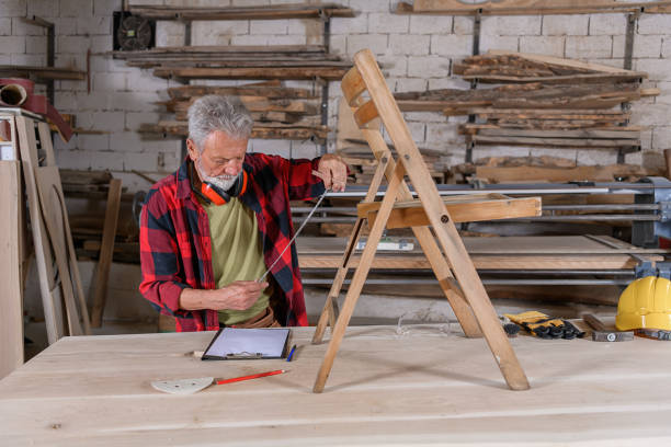 Professional Carpenter is Refinishing Wood Surface of an Old Wooden Chair While Using a Sand Paper and Other Carpentry Tools in a Large Workshop.
