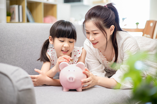 smiling asian mother and daughter saving money and putting coins into piggy bank - lying on sofa