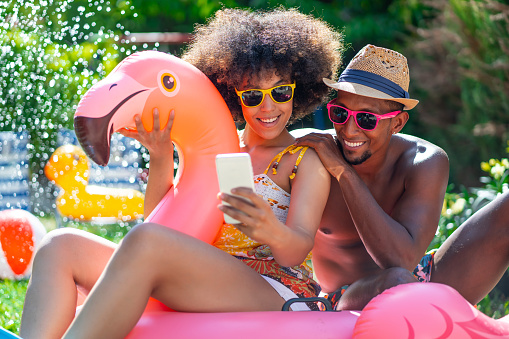 Afro Couple having staycation fun on back yard with inflatable pink flamingo and taking selfie, during COVID-19
