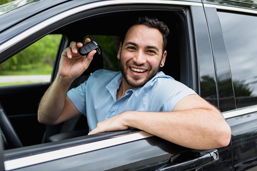 The mid adult man holds up his keys and smiles happily from driver's seat after buying his new car online.