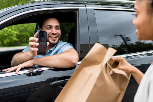 The mid adult man sits in his car and uses a mobile app to confirm his food order.  The mid adult woman smiles as she hands him his order.