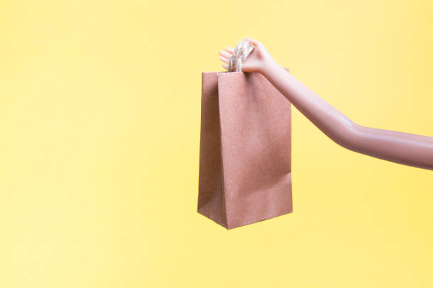 istock doll hand holds small brown paper bag, yellow background, copy space 1318010205