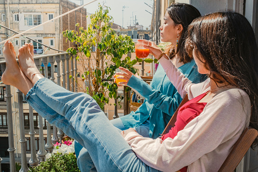 Two teenage girls , with natural juices, sunbathing in the morning, sitting on chairs on the balcony of a neighborhood apartment in Barcelona