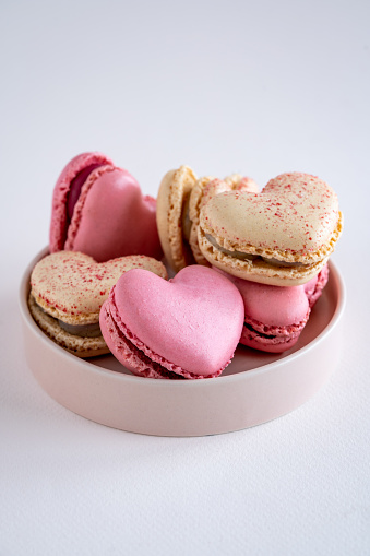 A group of heart-shaped French macaron cookies on a pink round plate, vanilla and strawberry flavour, copy space on neutral background, vertical