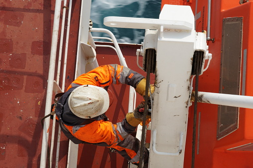 Seattle, United States - 12 13 2020:  Close up view on crew member with orange jacket and white helmet secured with harness to metal railing checking aft hook on life boat fixed in container vessel.
