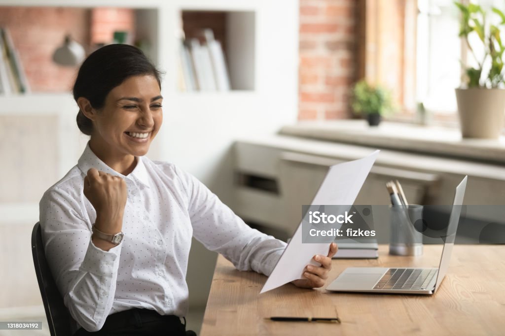 Overjoyed smiling Indian businesswoman reading good news in letter Overjoyed smiling Indian businesswoman reading good news in letter, sitting at work desk, excited young woman received scholarship, job promotion or offer, showing yes gesture, celebrating success Scholarship Stock Photo