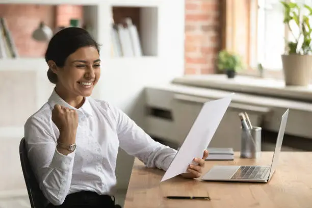 Overjoyed smiling Indian businesswoman reading good news in letter, sitting at work desk, excited young woman received scholarship, job promotion or offer, showing yes gesture, celebrating success