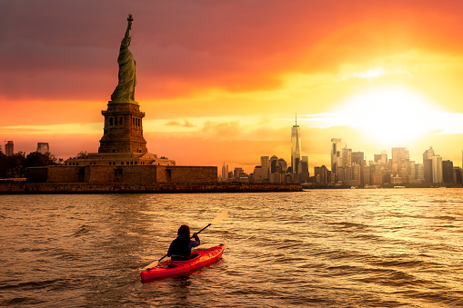 Adventurous Woman Sea Kayaking near the Statue of Liberty. Colorful Sunrise sky Art Render. Taken in Jersey City, New Jersey, United States.