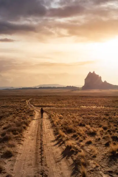 Woman walking on a dirt road in the dry desert with a mountain peak in the background. Dramatic Sunset Sky Art Render. Taken at Shiprock, New Mexico, United States.