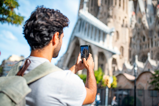 Over the shoulder view of 30 year old Hispanic male backpacker photographing Barcelona landmark with smart phone.