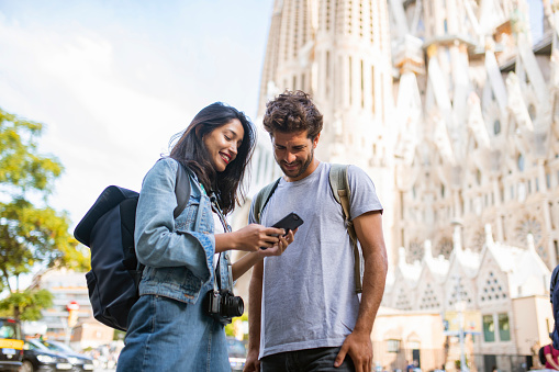 Low angle view of couple in 20s and 30s sightseeing in Barcelona and smiling as they look at smart phone with Sagrada Familia in background.