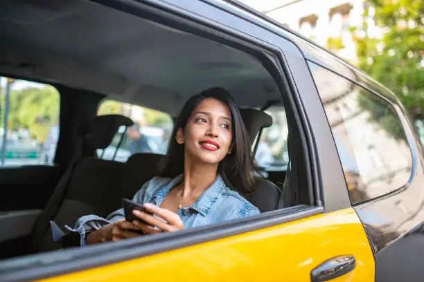 Side view of Indian female vacationer in mid 20s sitting with smart phone in backseat of taxi as she travels through downtown Barcelona.