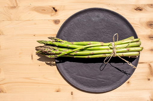 Healthy eating and lifestyle concept: On right frame a black plate with a bunch of fresh and organic asparagus on a wooden table. Preparing for cooking. Ingredients for a traditional recipe. Natural food background with large copy space.