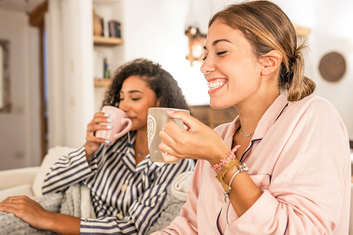 Couple of cohabiting girls in pajamas relaxing under blanket on cozy sofa drink a cup of tea while having fun talking and joking with each other. Real life moments of diverse multiracial gay couple