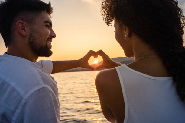 istock Young just married multiracial couple in love making heart shape with hands with setting sun in centre of it, looking in the eyes smiling. Romantic scene of Hispanic girl and boyfriend view from back 1318089206