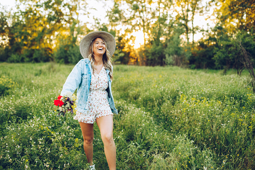 Beautiful happy blonde young woman holding wild flowers and hat enjoying sunny day in nature, feminine freedom