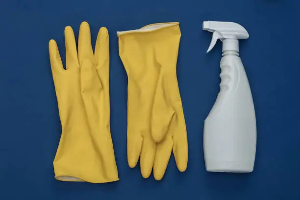 Photo of Yellow rubber gloves and bottle spray of detergent on classic blue background. Color 2020. House cleaning theme. Top view