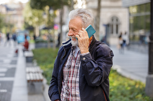 An Older Man is Walking in the City Streets and Talking with his Friends over the Smart Phone. Happy Successful Business Person is Walking in the City Streets to the Office while Using a Phone.