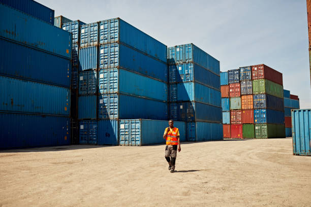 Full length front view of Black man in early 40s wearing reflective vest and talking on two-way radio as he approaches camera amidst shipping containers.