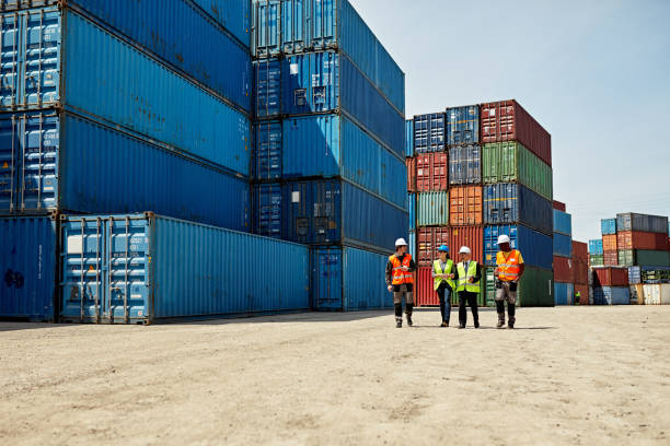 Logistics Team Walking Together in Inland Port Mid distance view of diverse cargo handlers and supervisors approaching camera in reflective vests and hardhats amidst stacks of cargo containers. delivering stock pictures, royalty-free photos & images