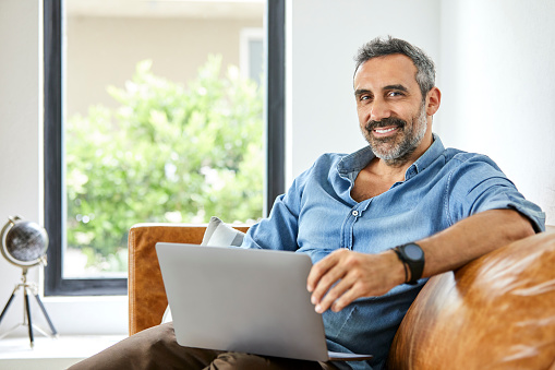 Portrait of gay man sitting on sofa. Smiling mature man is using laptop. He is in casuals at home.