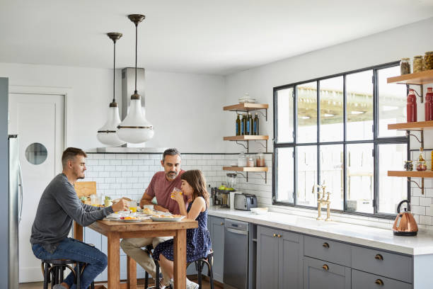 Girl having breakfast with parents at home Girl eating breakfast with parents. Homosexual couple is having food with daughter. They are sitting in kitchen. gay dinner table stock pictures, royalty-free photos & images