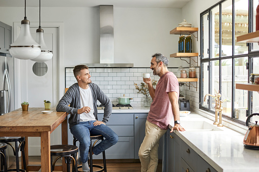 Happy homosexual couple talking at home. Smiling gay men are having coffee. They are in kitchen.