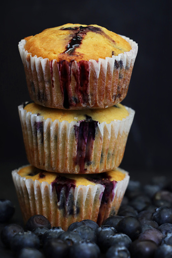 Stock photo showing close-up view of fresh blueberries surrounding a pile homemade blueberry muffins in paper cake cases against a dark background.