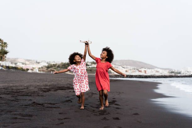 hermanas gemelas negras corriendo en la playa mientras juegan con un avión de juguete de madera - concéntrate en las caras - afrocaribeño fotografías e imágenes de stock