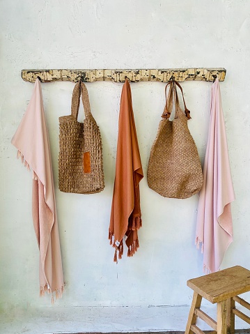 Vertical rustic hanging rack coat hooks with pale coral pink and burnt terracotta orange Turkish organic cotton beach towels, hand woven beach bags and wood stool in hallway entrance to summer beach house
