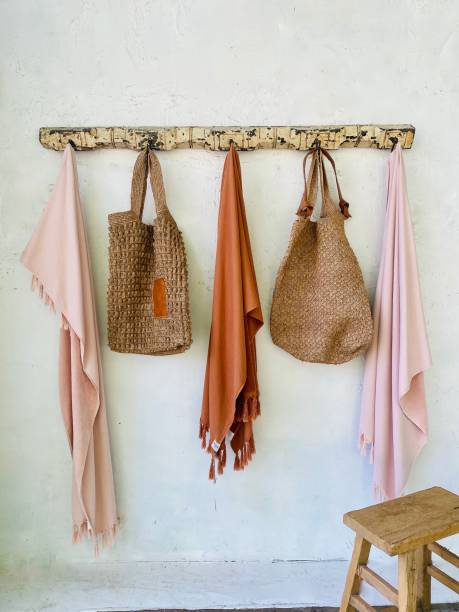 Wall Hooks Holding Beach Wear in Summer House Hallway Vertical rustic hanging rack coat hooks with pale coral pink and burnt terracotta orange Turkish organic cotton beach towels, hand woven beach bags and wood stool in hallway entrance to summer beach house beach bag stock pictures, royalty-free photos & images