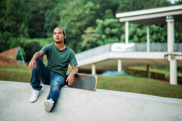 istock Asian male skateboarder taking a break and looking away in a skate park 1318138607