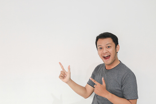 Wow and surprised face with smile of young Asian man in gray t-shirt with hand point on empty space.