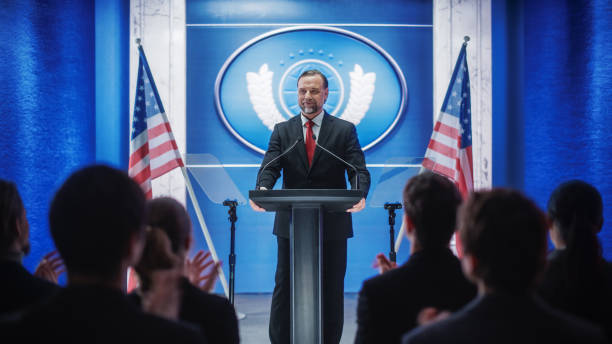 istock Charismatic Organization Representative Speaking at a Press Conference in Government Building. Press Officer Delivering a Speech at a Summit. Minister at Congress. Backdrop with American Flags. 1318165794