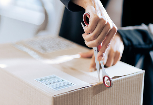 Hand of a female opening a cardboard box seal with scissor. Woman opening her courier parcel.