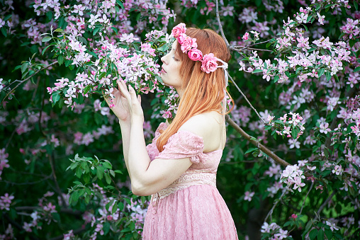 A young woman in a pink lace dress and a wreath of roses inhales the fragrance of a flowering tree. An idea for a photo shoot.