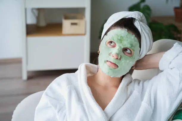 Photo of Beautiful young woman applies moisturizing green funny bubble face mask.Lying on couch. White bathrobe,towel.Self skin care,anti age morning treatment for wrinkles at home.Apartment with green plants