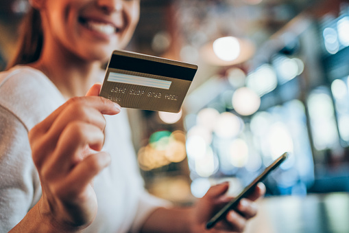 Portrait of young woman doing shopping online with smartphone and payment by credit card. Woman holding smartphone and credit card in urban cafe. Focus is on the card.