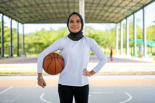 Beautiful Young Asian Female Athlete wearing a sports hijab posing at an outdoor public basketball court as a basketball player.