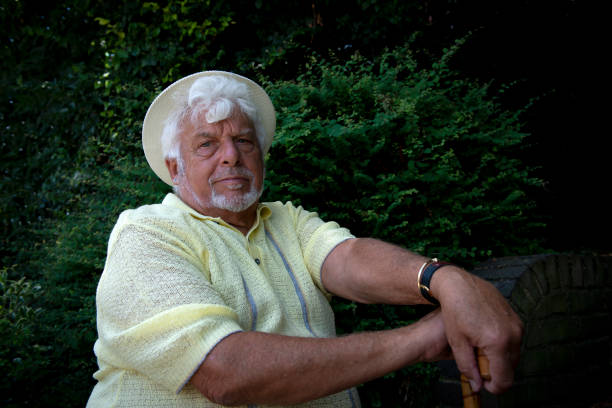 A portrait of over 70 years old English man. The man is wearing a yellow hat and yellow a short. He is looking at the camera. His hands are on the his walking stick.