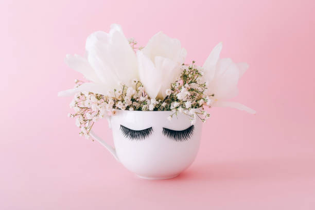 Creative white mug of flowers with long eyelashes on pink background.