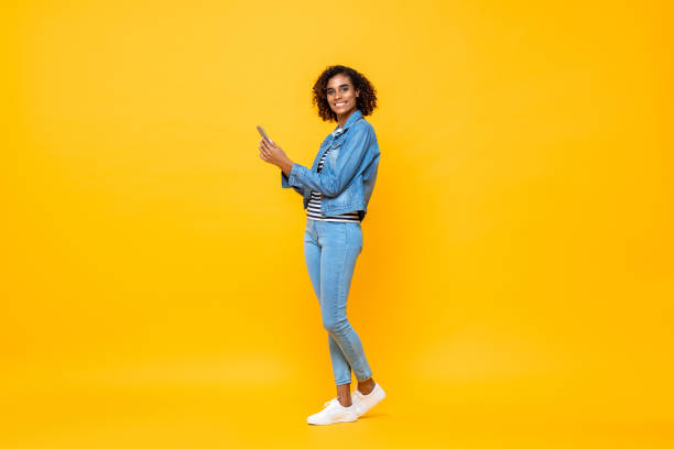 verticale pleine longueur de jeune femme afro-américaine de sourire regardant l’appareil-photo tout en retenant le téléphone portable dans le fond jaune d’atelier d’isolement - cadrage en pied photos et images de collection